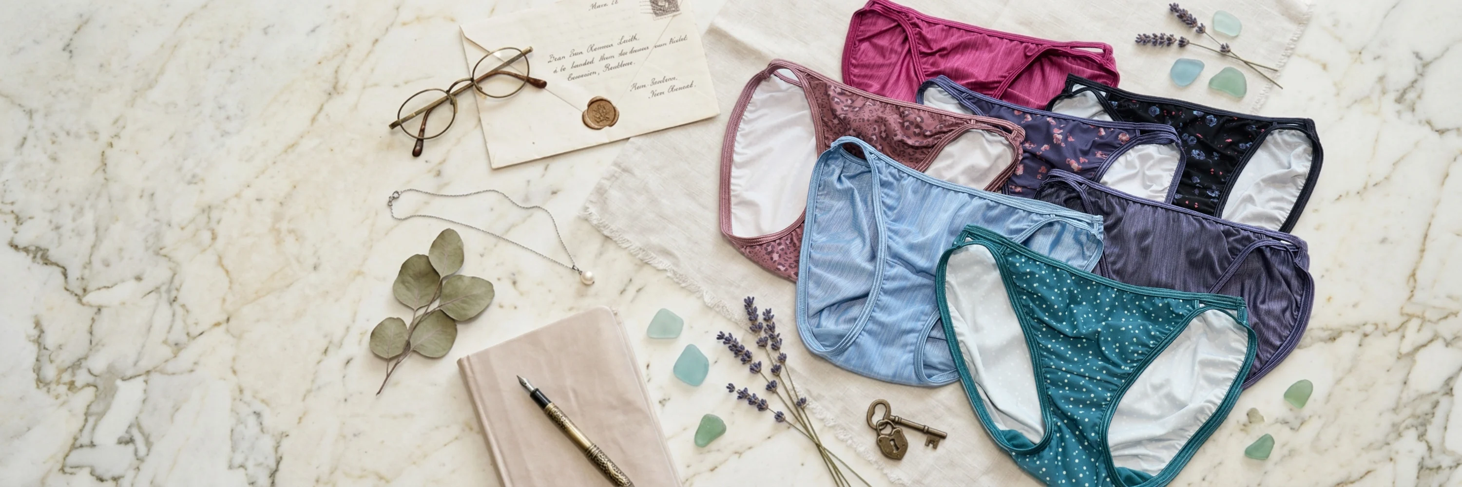 Assorted Vanity Fair panties in solid colors and prints displayed on a light marble tabletop alongside eyeglasses, a journal, jewelry, lavender stems, and decorative accents.