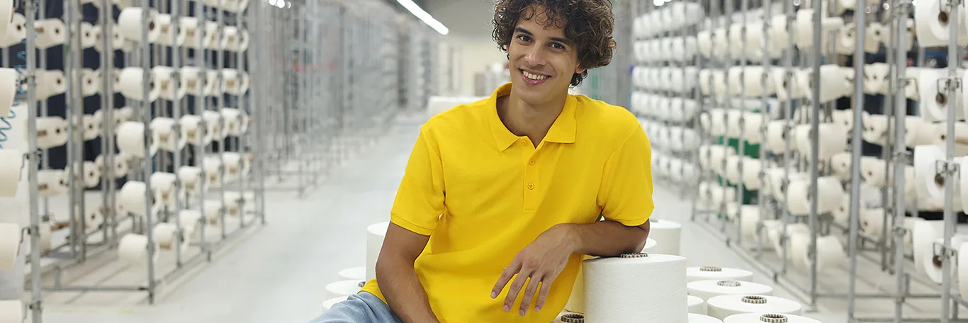 A man sits in a yarn storage area within a production facility, surrounded by shelves filled with spools of yarn.