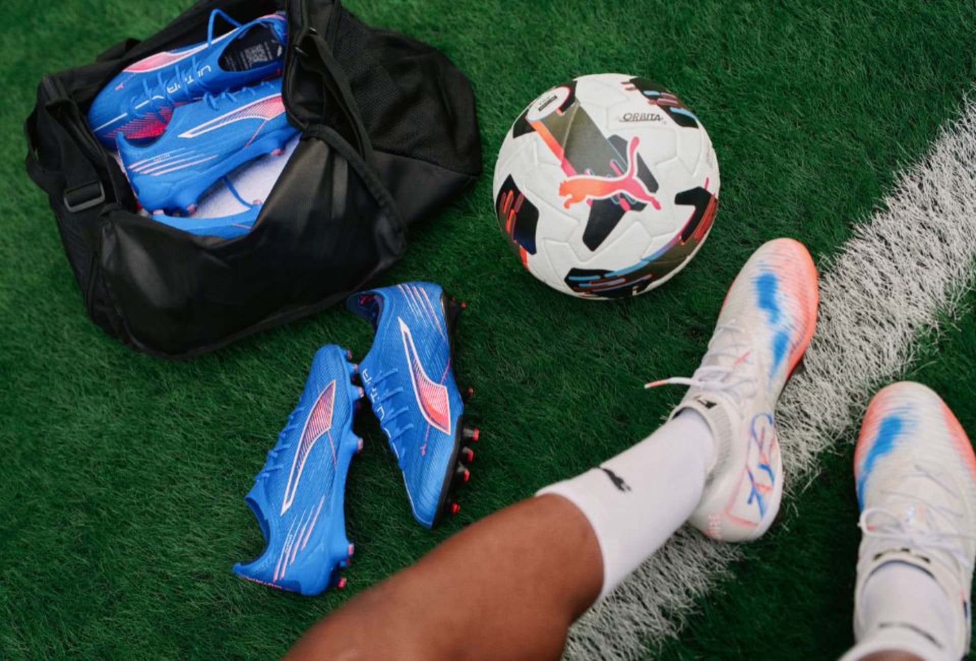 Fussballschuhe und eine Sporttasche gefüllt mit Fussballschuhen auf einem Rasenfeld.