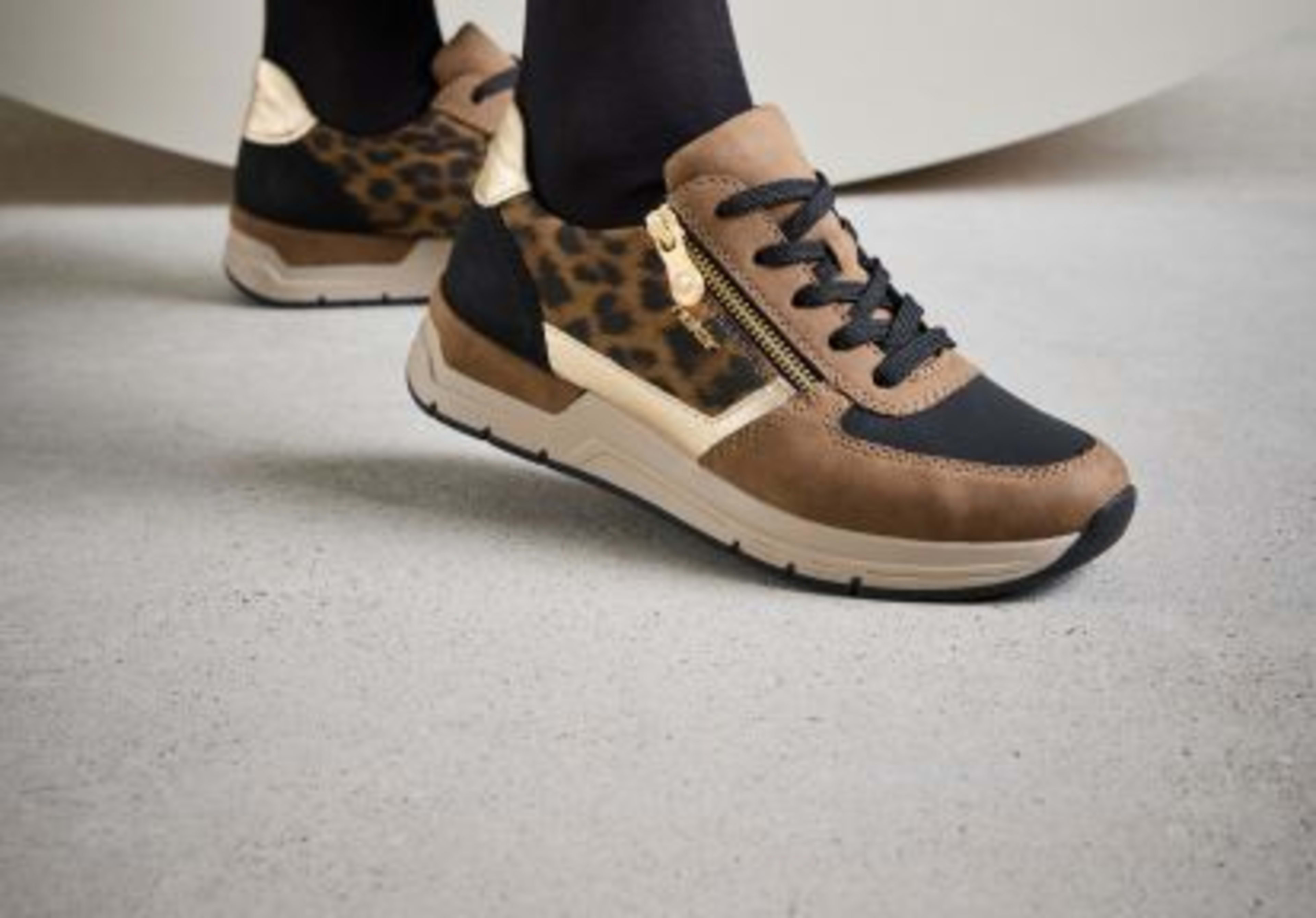 Rieker Damen-Sneaker in Braun mit Leo-Print und goldenen Details, kombiniert mit schwarzen Socken und Hose.