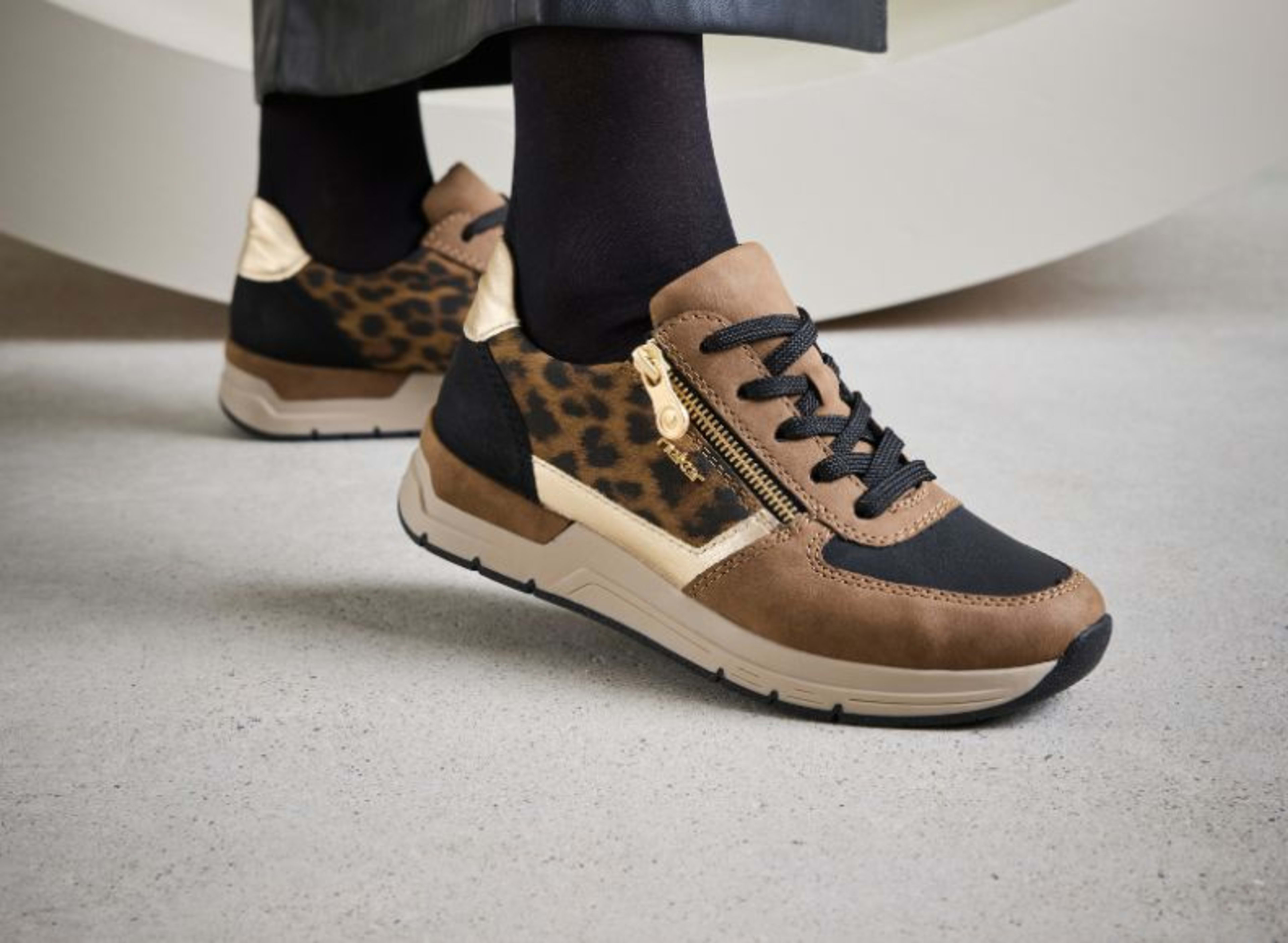 Rieker Damen-Sneaker in Braun mit Leo-Print und goldenen Details, kombiniert mit schwarzen Socken und Hose.