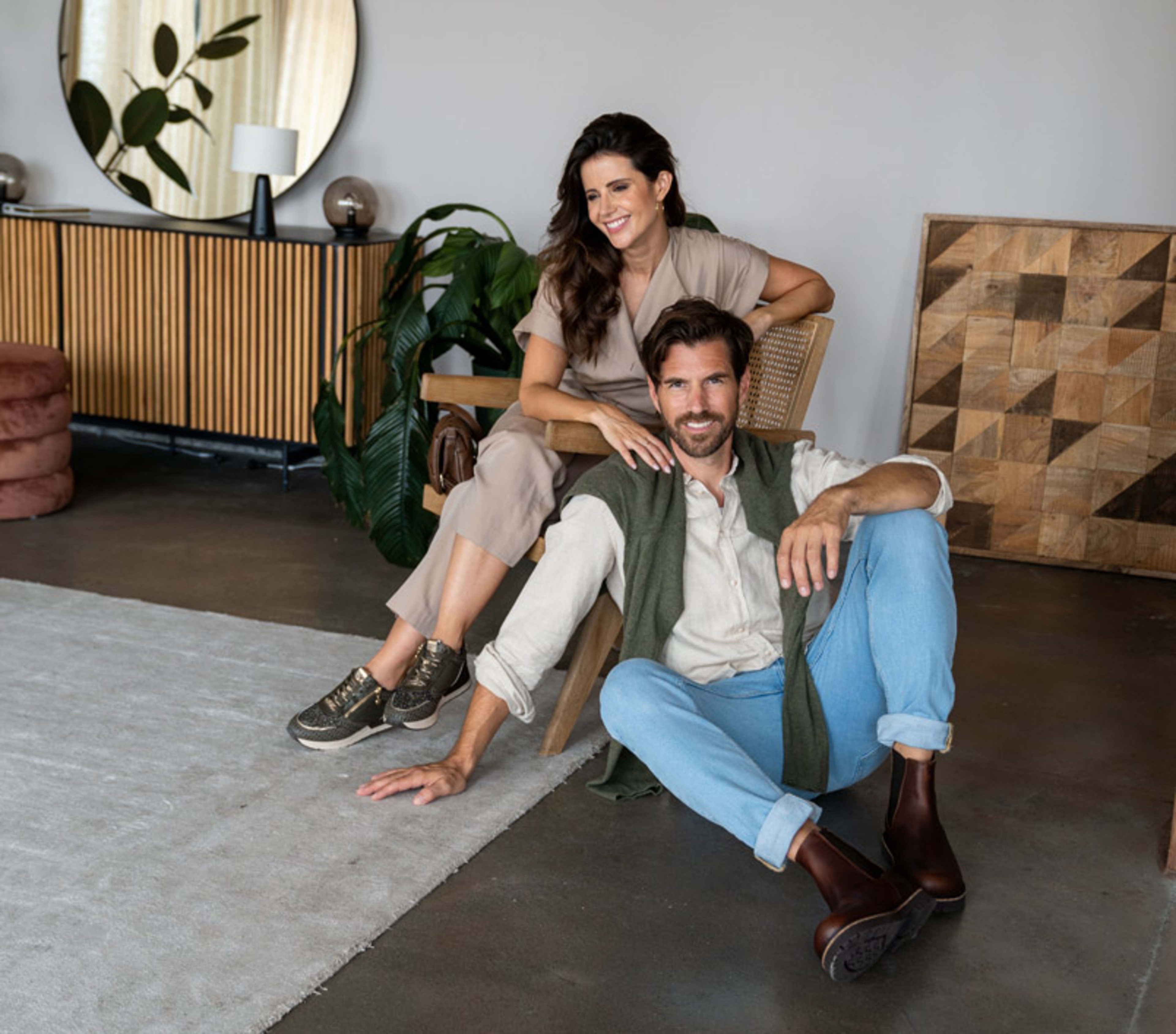 Eine Frau mit weißem Kleid und orangenen Sandaletten sitzt mit einem Mann auf einer Bank. Sie trinkt ein orangenes Getränk und er einen Kaffee. Der Mann trägt Espadrilles, eine lange graue Hose und ein langärmliges beiges Oberteil.