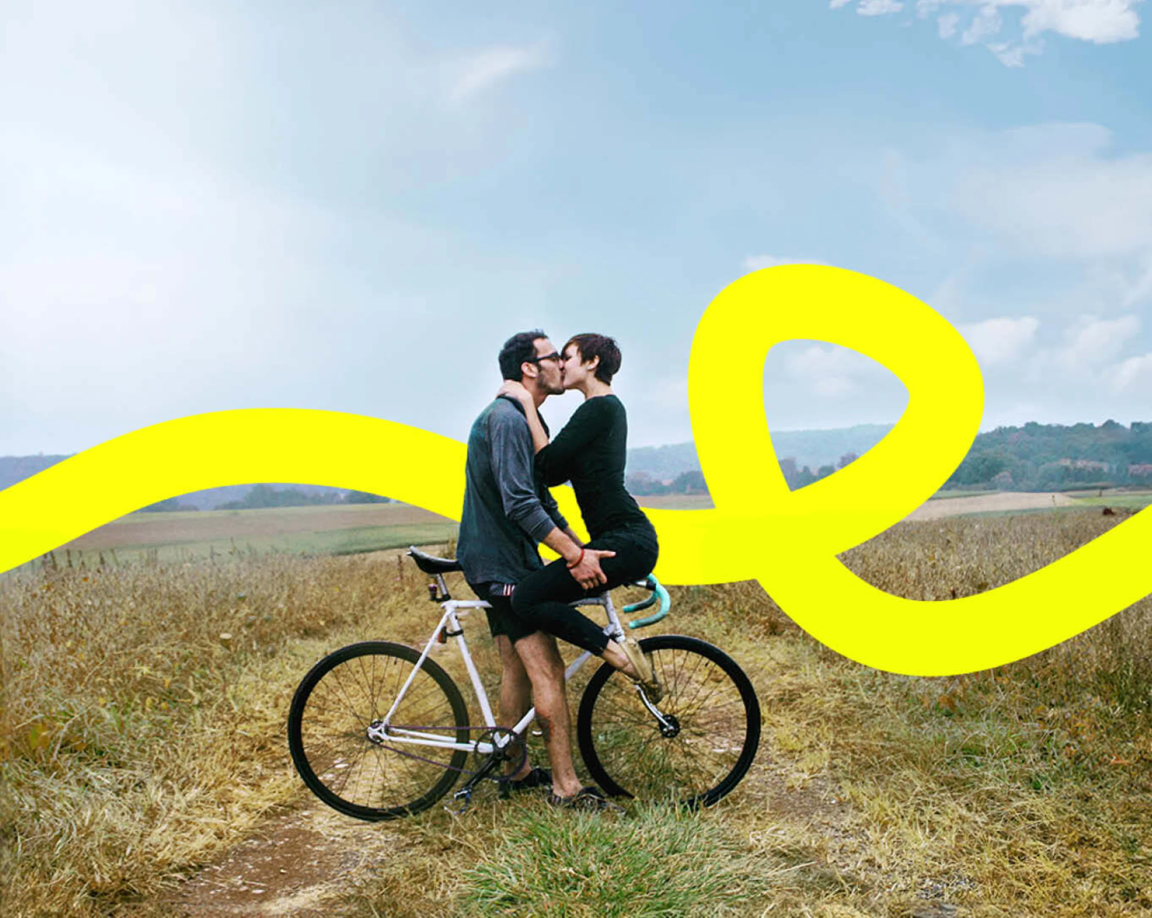 Eine Frau und ein Mann küssen sich auf einem Fahrrad vor einer Wiese.