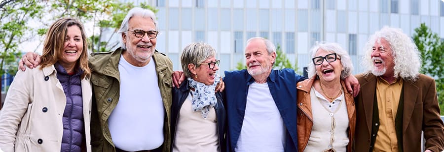 Grupa sześciu szczęśliwych starszych turystów i turystek stoi razem, obejmując się i uśmiechając na tle nowoczesnego budynku