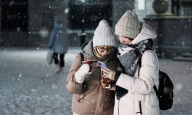 Dwie osoby w zimowych czapkach i kurtkach patrzą na smartfona podczas opadów śniegu w mieście - używanie internetu za granicą na feriach zimowych z eSIM