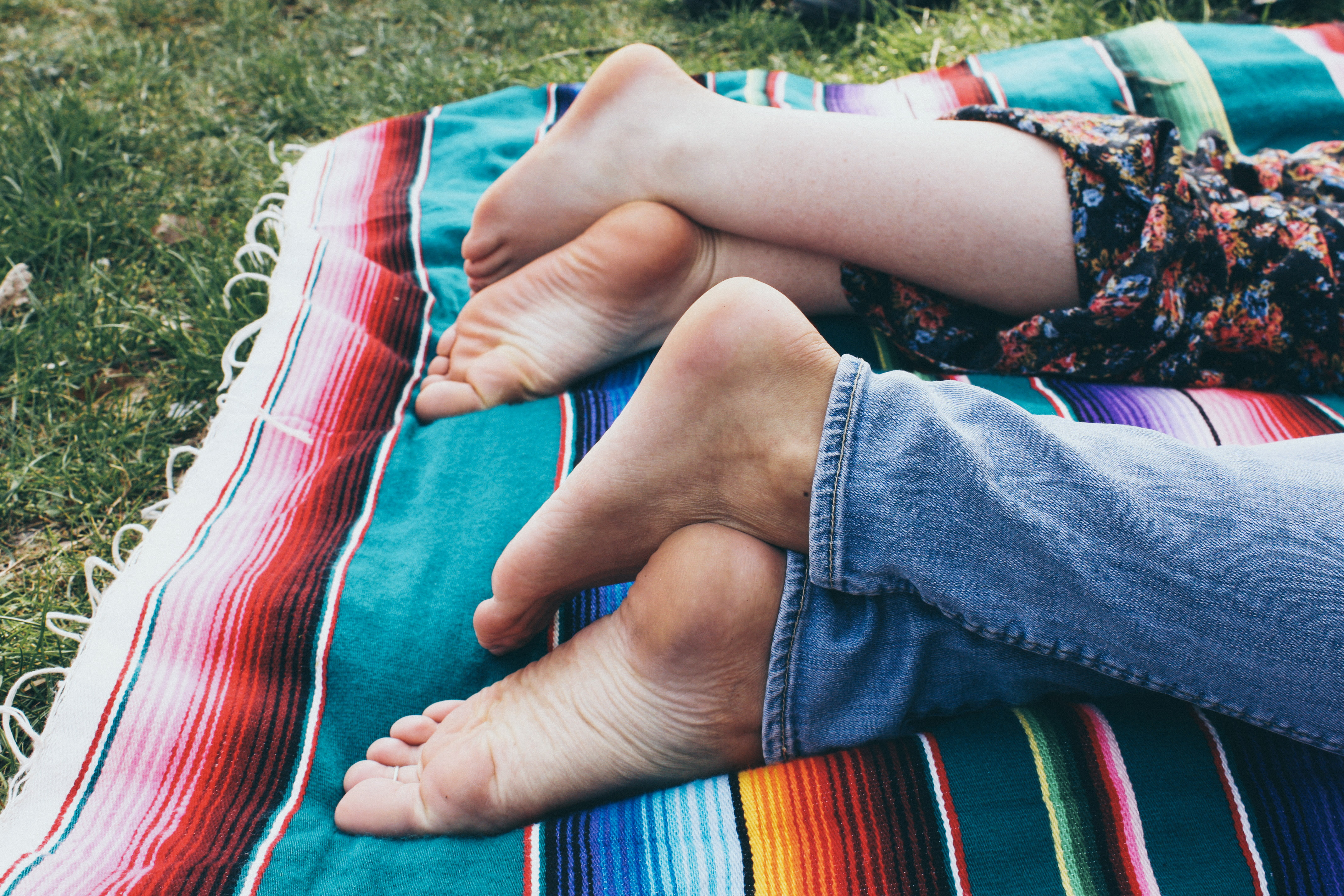 two women's feet laying on a rainbow blanket in a park 