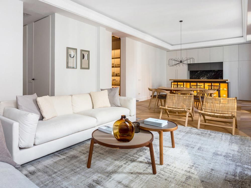 A bright, minimalist living room with a white sofa, wooden coffee tables, and a dining area.