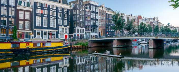Traditional Dutch canal houses reflected in calm water with yellow houseboat and stone bridge in Amsterdam
