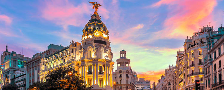 Historic ornate building with dome and winged statue illuminated at sunset in Madrid's Gran Via with colorful sky