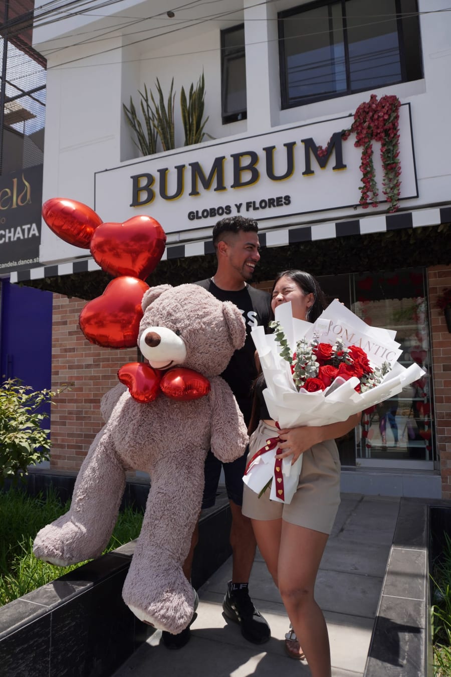 Pareja con globos de corazón y un peluche frente a la tienda Bumbum