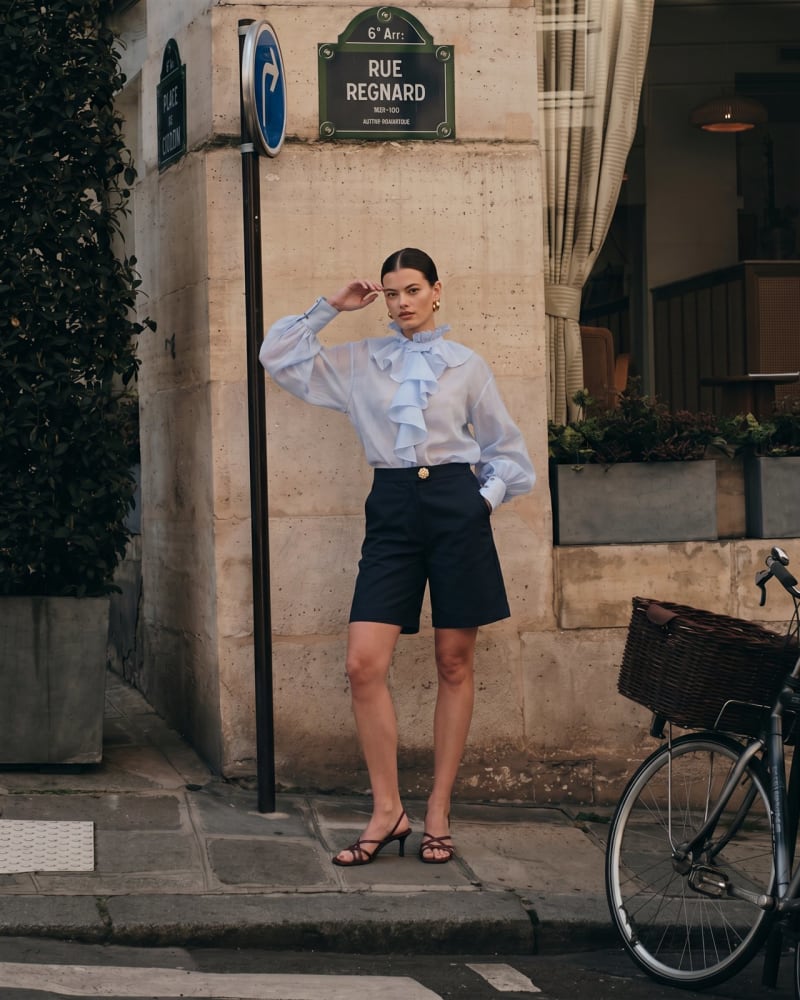 Look de bermudas navy con blusa de volantes transparente, fotografía ecommerce con modelo IA