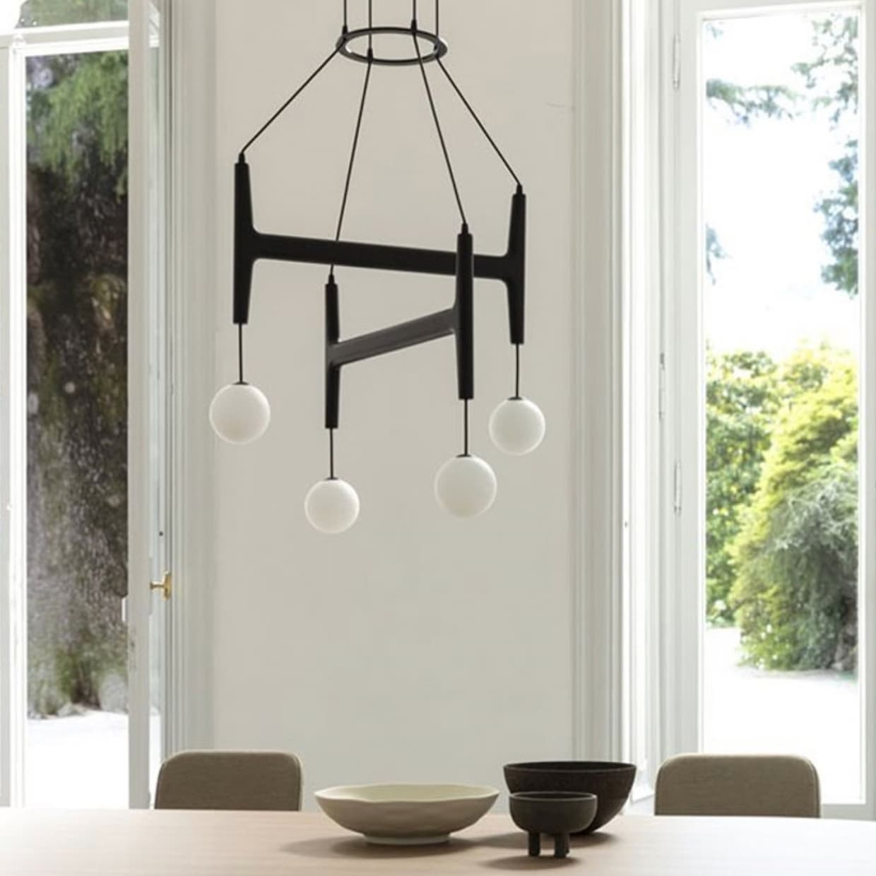 Dining room, large open windows overlooking the garden. Wooden table, vases and suspended luminaire with glass globes.