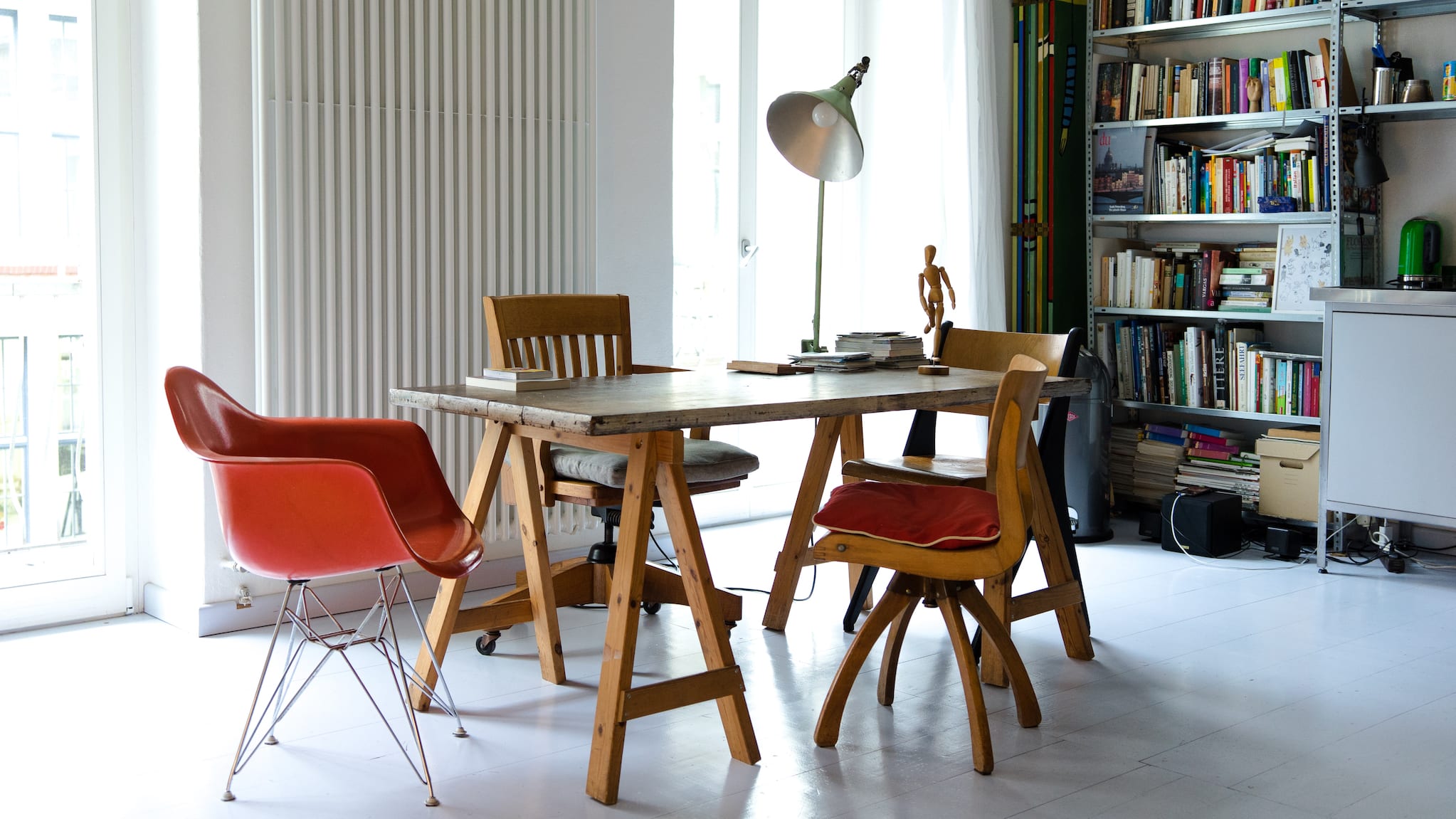 Loft with large windows, table on trestles, chairs of various styles and metal shelves filled with books.