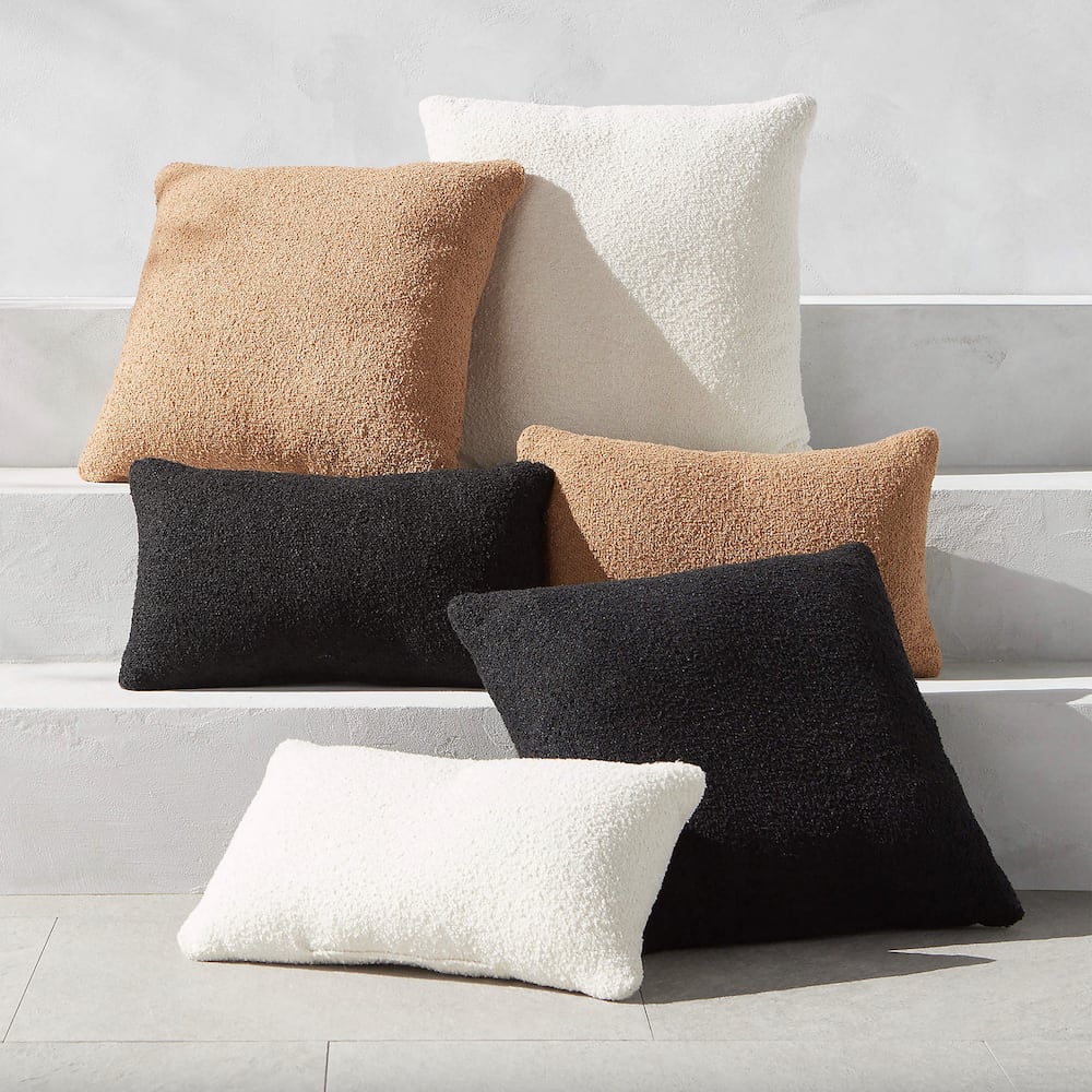 Coussins carrés et rectangulaires en laine bouclée blanc, noir, camel, disposés dans des marches blanches ensoleillées