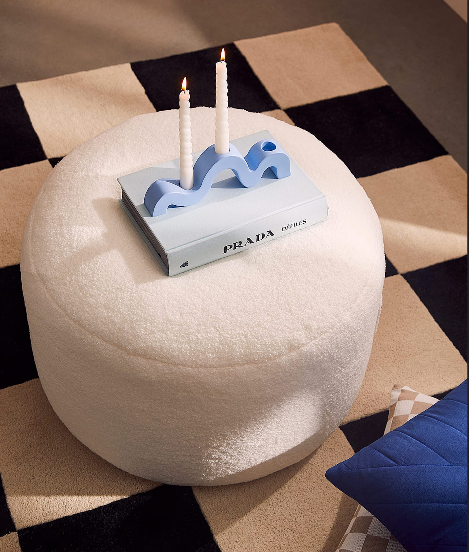 Pouf en laine bouclée blanc ivoire, livre Prada, bougies torsadées blanches, tapis damier noir et blanc