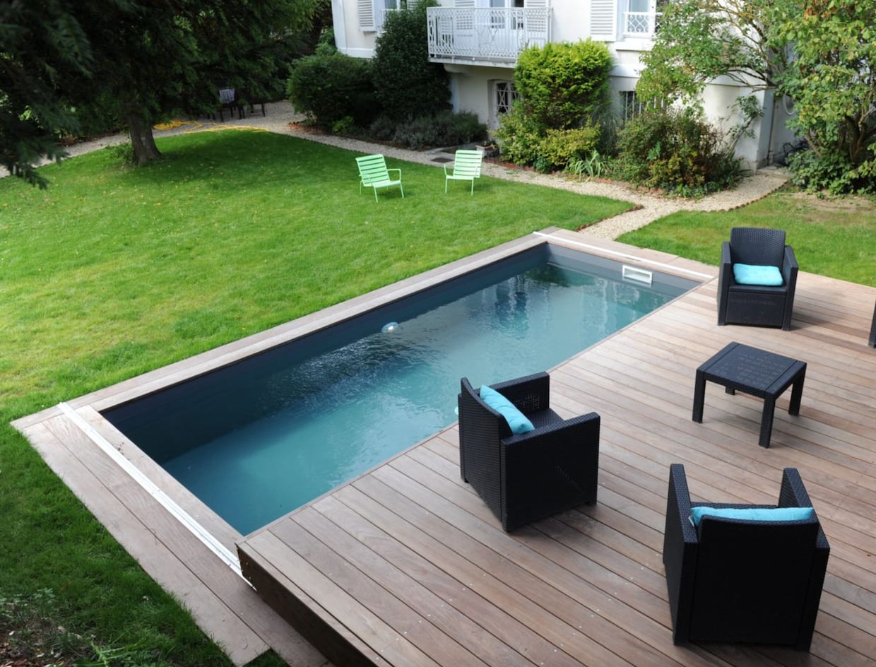 Belles piscines terrasse coulissante en lattes de bois, fauteuils et table en vannerie noire, coussins turquoise
