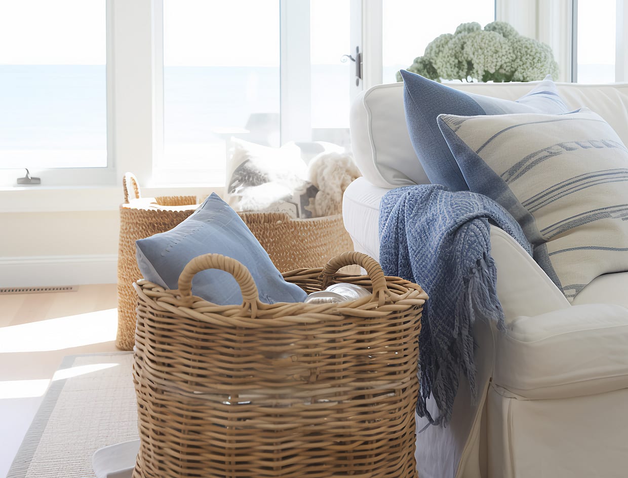 Déco voyage, salon fenêtre sur océan, avec canapé crème, coussins rayés bleu ciel et blanc, paniers tressés au sol et jeté
