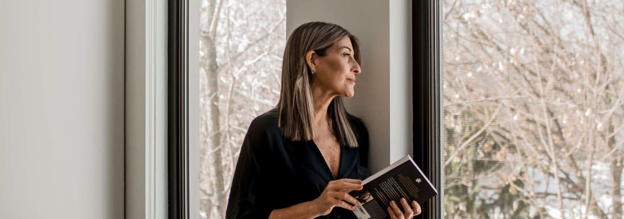 Une femme appuyée sur le mur de sa maison qui regarde par la fenêtre. Elle tient un livre dans ses mains
