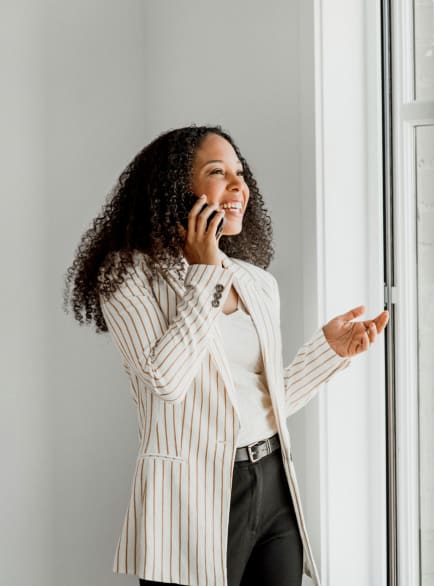 Une femme qui sourit en parlant au téléphone. Elle est debout près d'une fenêtre.