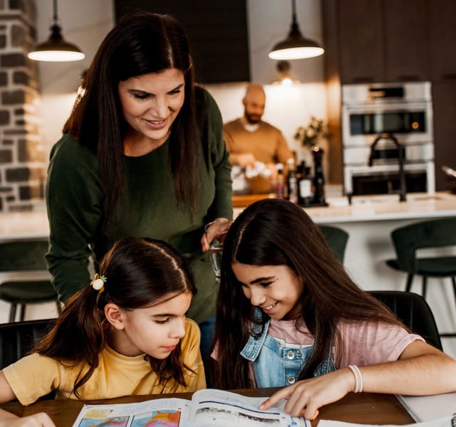 Deux enfants qui regardent un livre dans la cuisine. Leur mère est derrière eux et leur père cuisine
