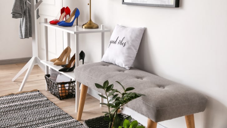 Petit hall d'entrée avec meuble blanc présentant des souliers et sur lequel repose une lampe, banc gris sur lequel s'asseoir, plante et tapis au sol