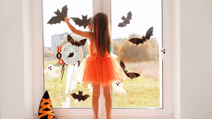 A young girl stands on a windowsill while sticking paper bat decorations on the window glass