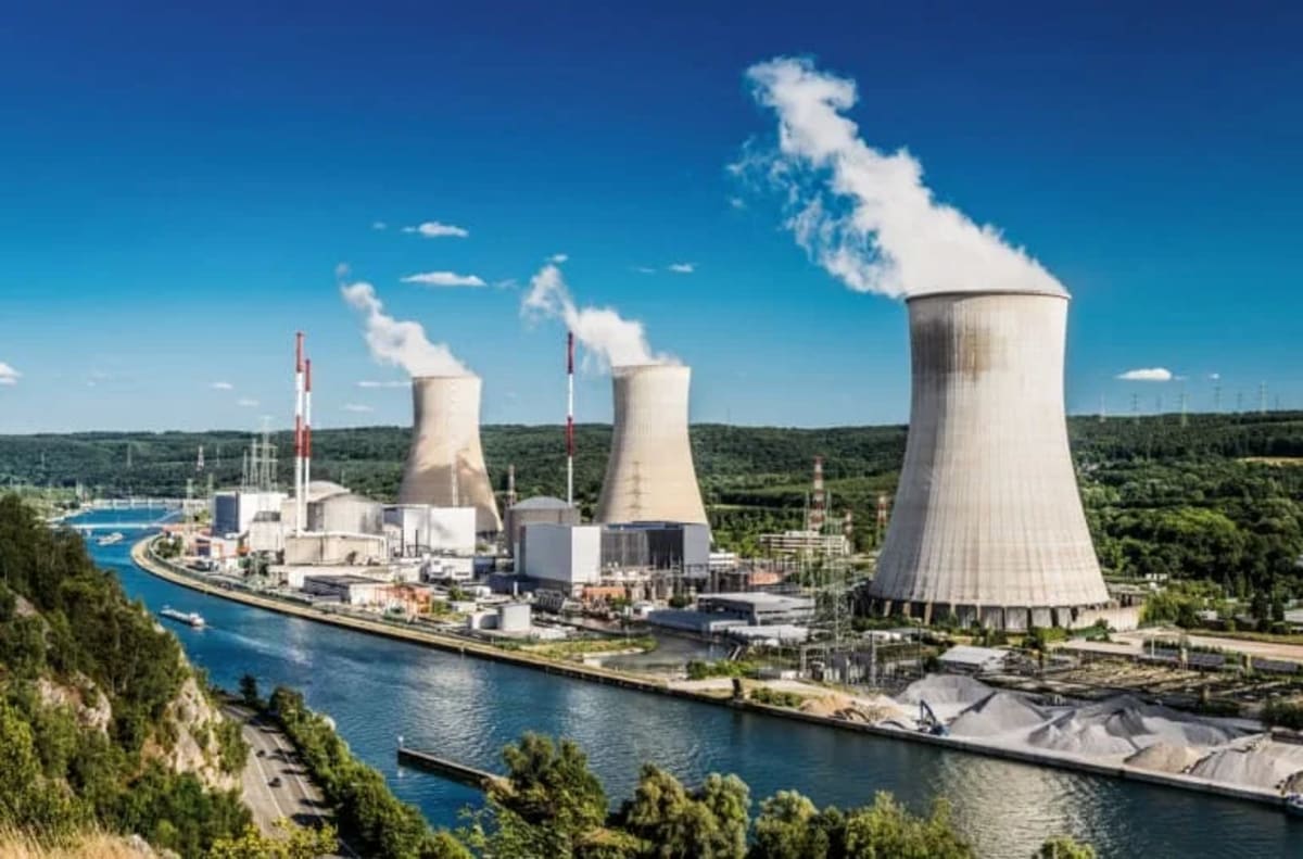 Vista de una planta nuclear moderna cerca de un río, con grandes torres de enfriamiento expulsando vapor bajo un cielo despejado