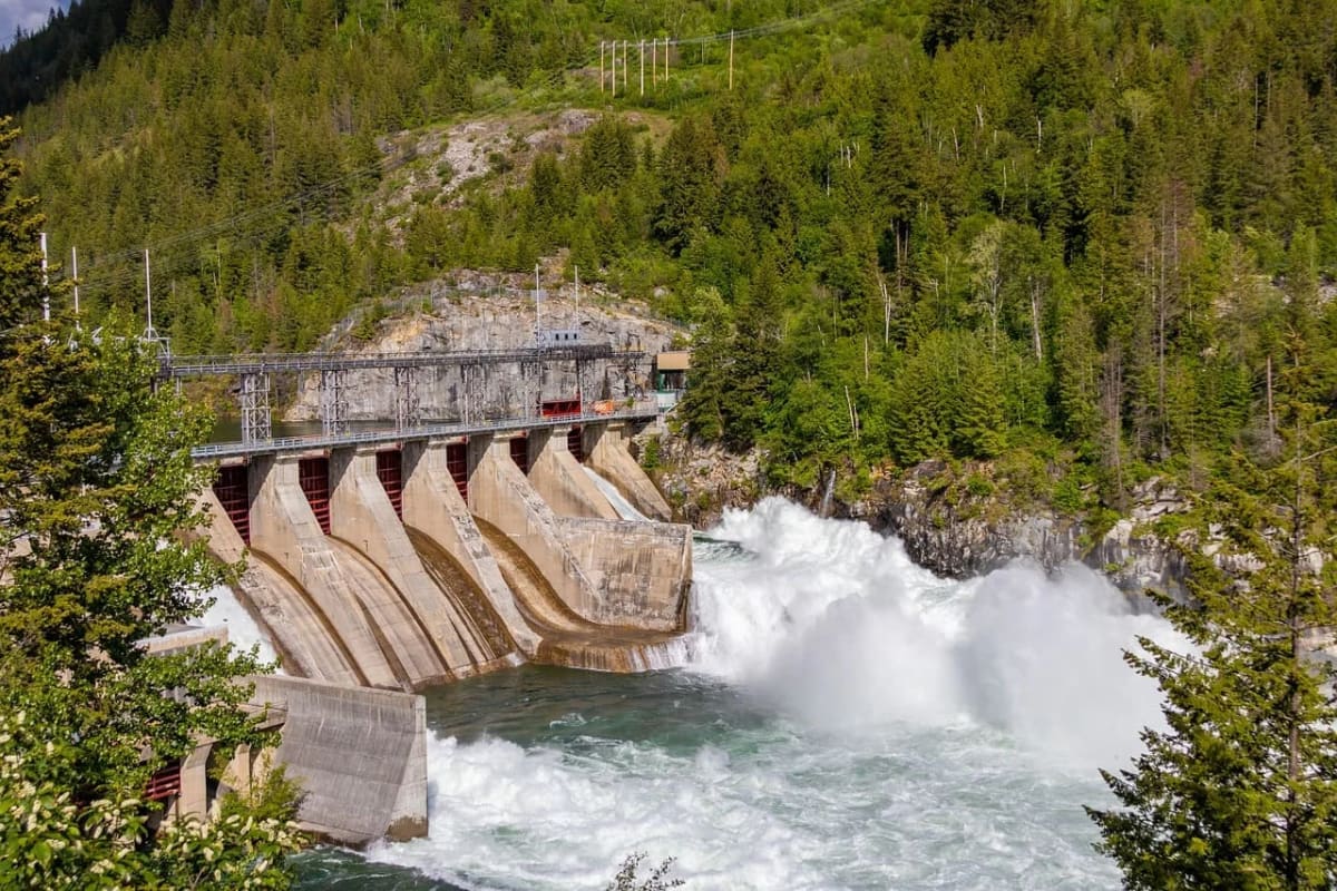 Central hidroeléctrica en un entorno natural, con agua fluyendo a gran velocidad a través de una presa rodeada de vegetación y montañas