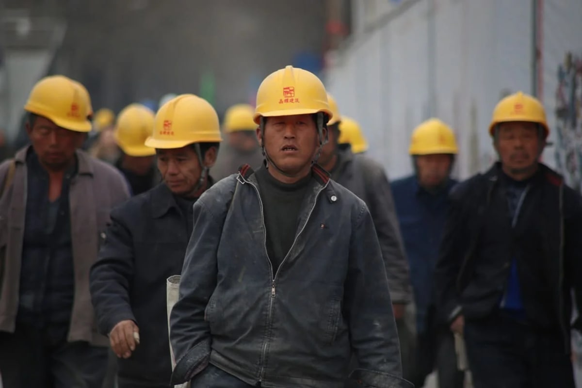 Grupo de trabajadores chinos de la construcción caminando, vistiendo cascos de seguridad amarillos y ropa de trabajo, en un entorno industrial.