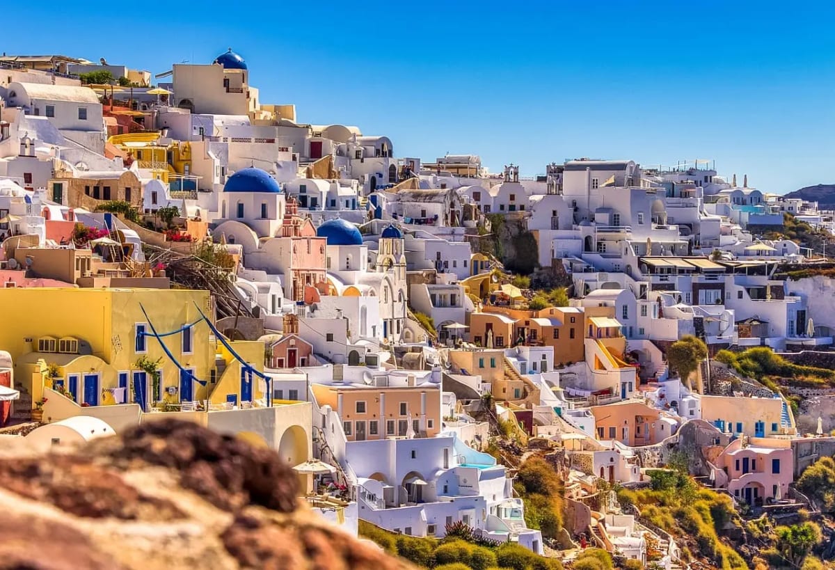 Vista panorámica de Santorini, Grecia, con casas blancas y cúpulas azules iluminadas por el sol bajo un cielo despejado.