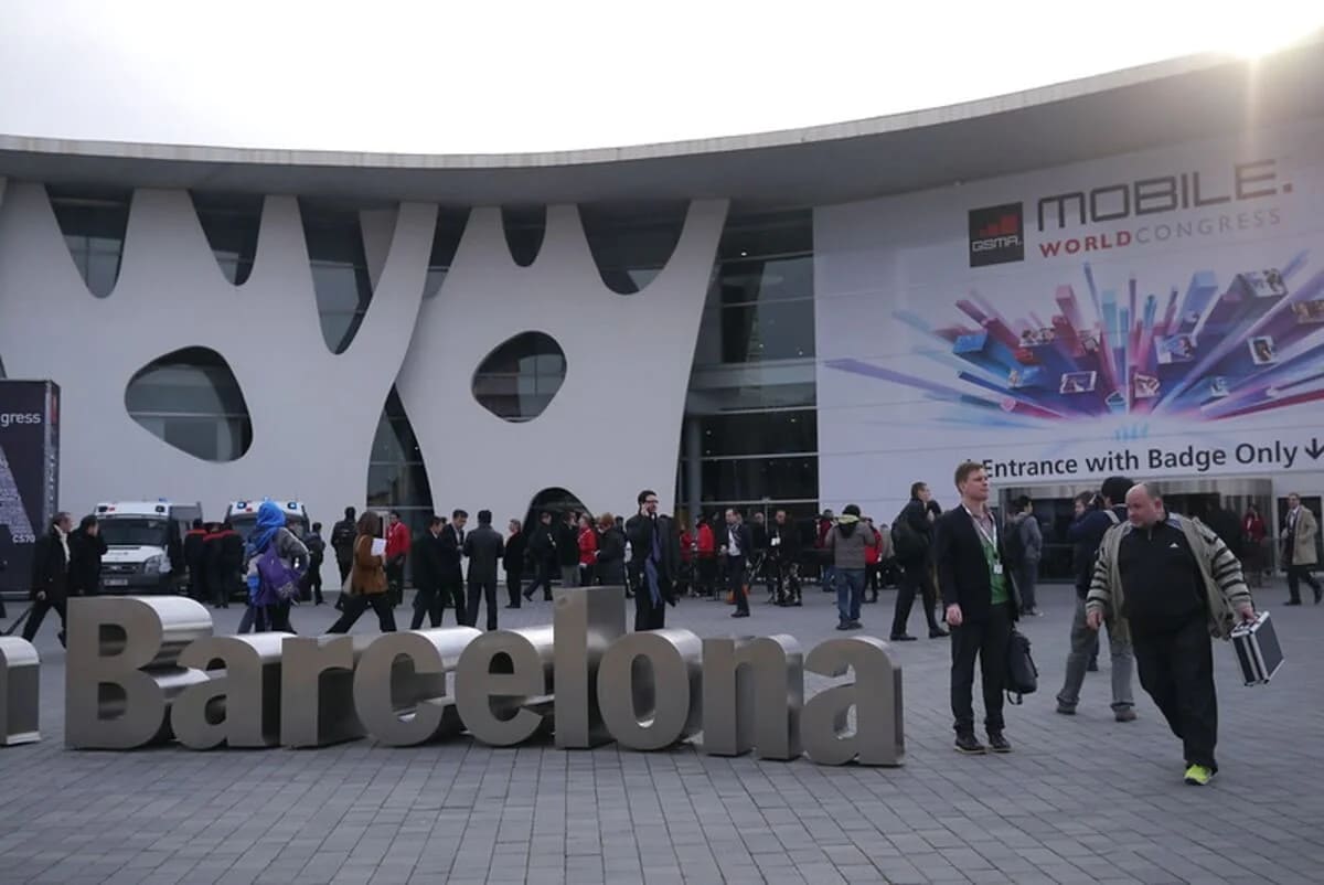 Entrada principal del Mobile World Congress en Barcelona, con asistentes caminando frente al edificio y un cartel del evento visible.