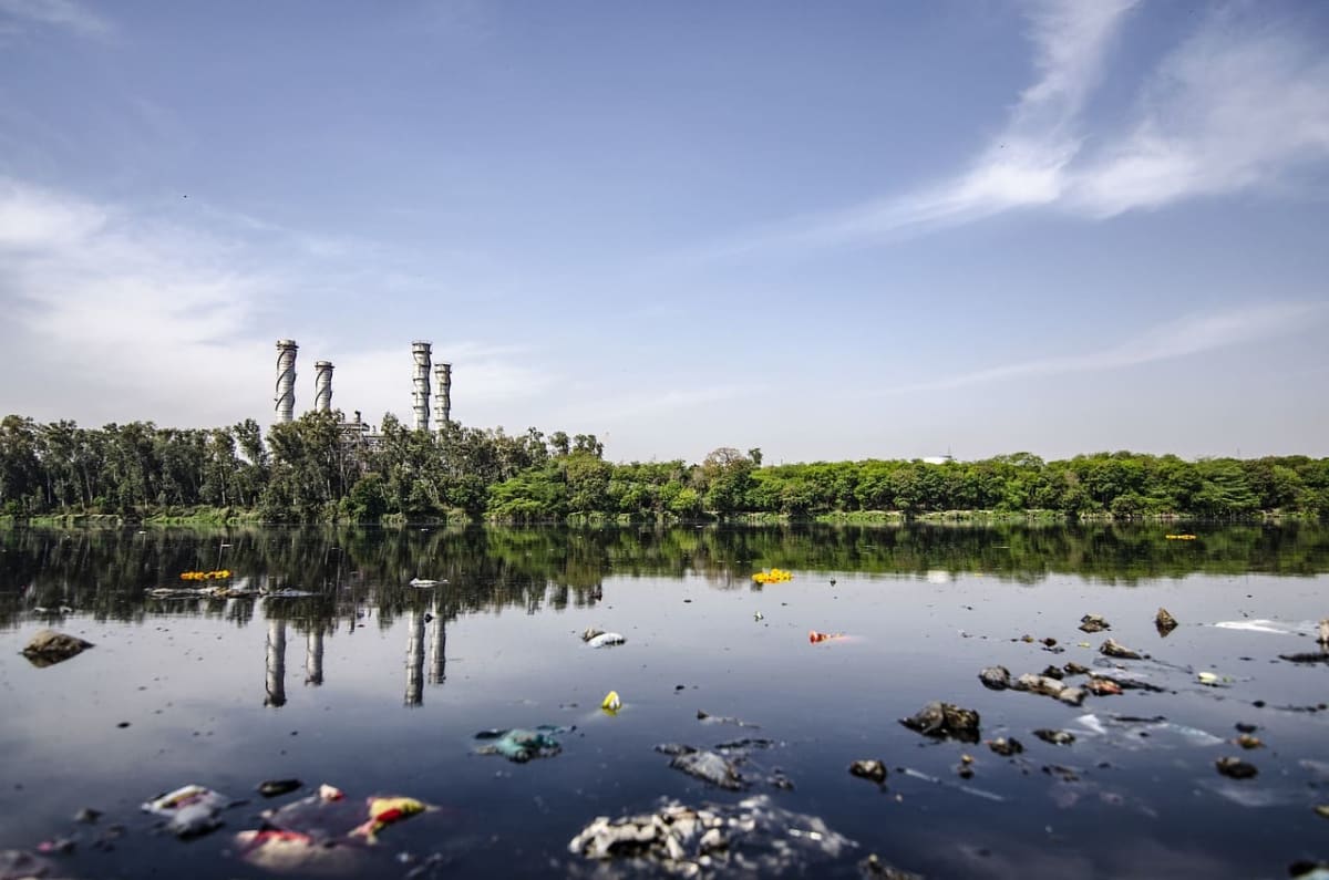 Contaminación visible en un río lleno de residuos sólidos, ubicado junto a una planta industrial rodeada de árboles frondosos.