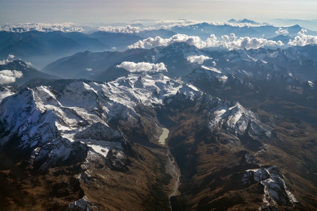 Vista aérea de la cordillera de los Alpes con cumbres cubiertas de nieve, valles profundos y bancos de nubes flotando.