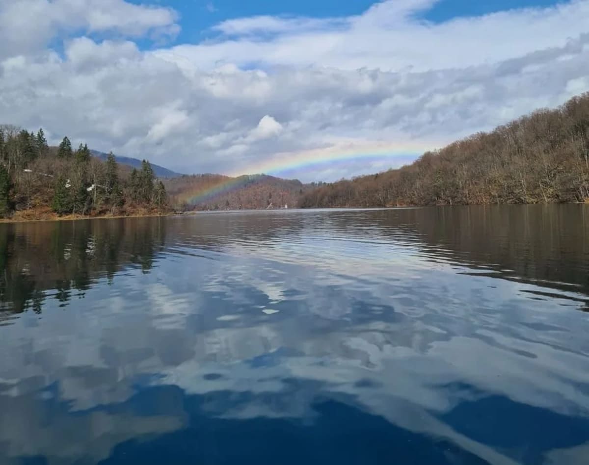 Fotografía invernal del lago Kozjak con un arcoíris en el horizonte, rodeado de colinas arboladas.