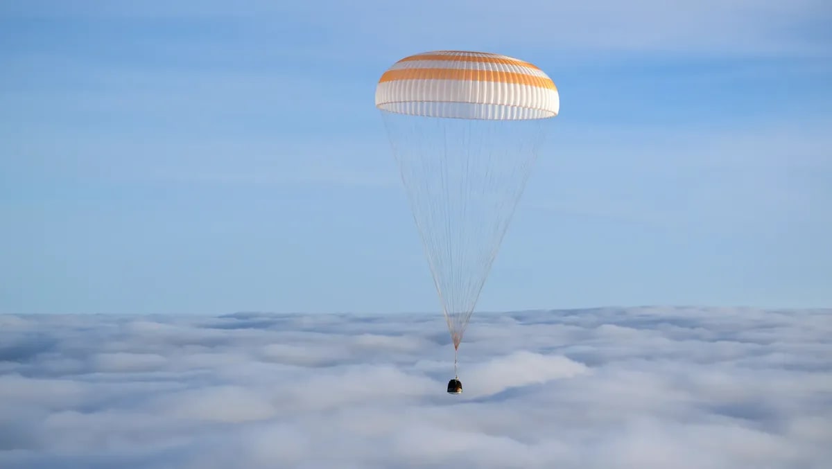 Cápsula Soyuz MS-26 descendiendo sobre una capa de nubes con su paracaídas desplegado.