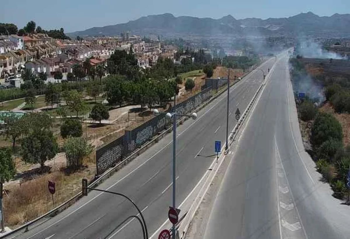 Vista aérea de una avenida en Málaga con humo por incendio en el margen derecho, cerca de zona residencial y montaña al fondo.