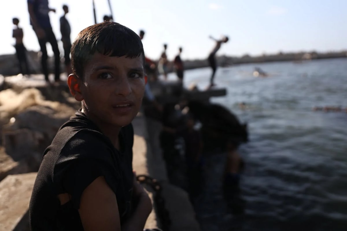 Niño en Gaza sentado junto al mar mientras otros niños juegan en el agua