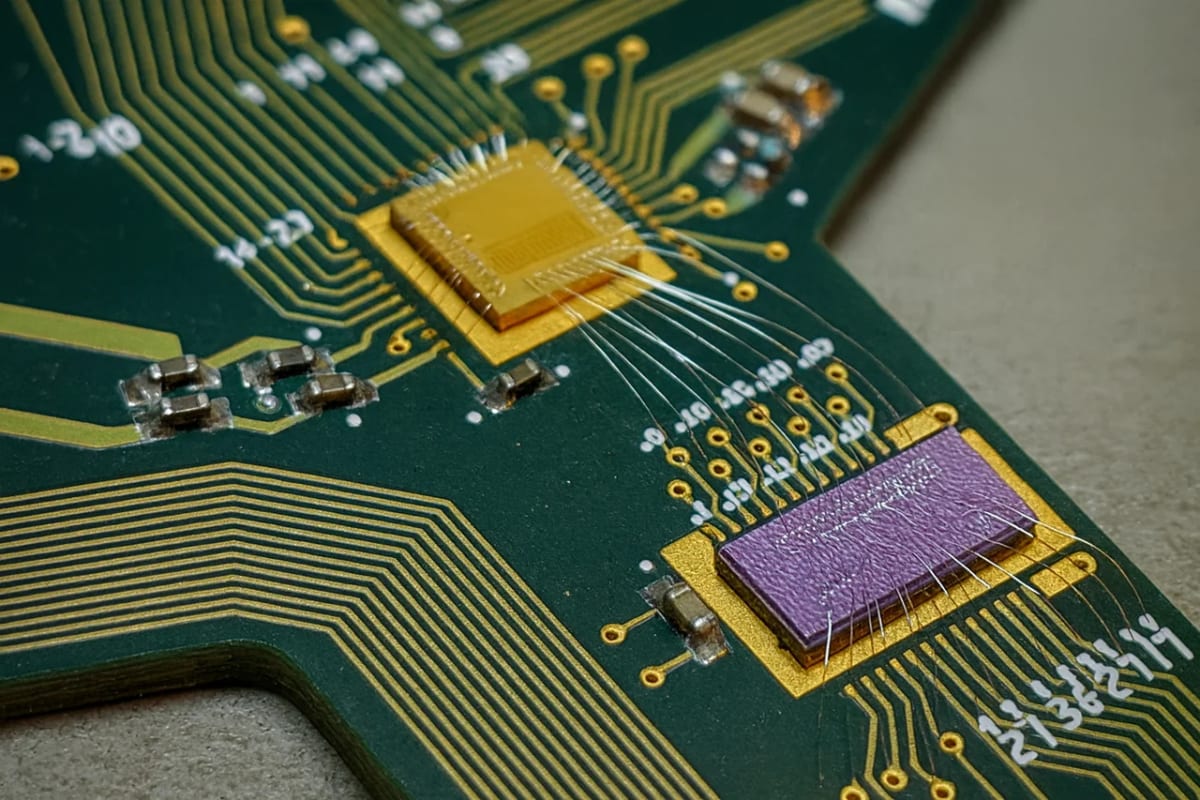 Fotografía macro de un chip fotónico montado sobre una placa verde, con microcables metálicos conectando los circuitos y trazas doradas visibles en detalle