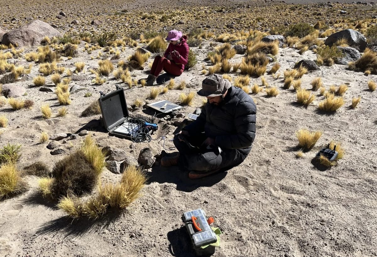 Investigadores realizan mantenimiento de un sismómetro en el desierto de Atacama, con un técnico en primer plano y una estudiante al fondo