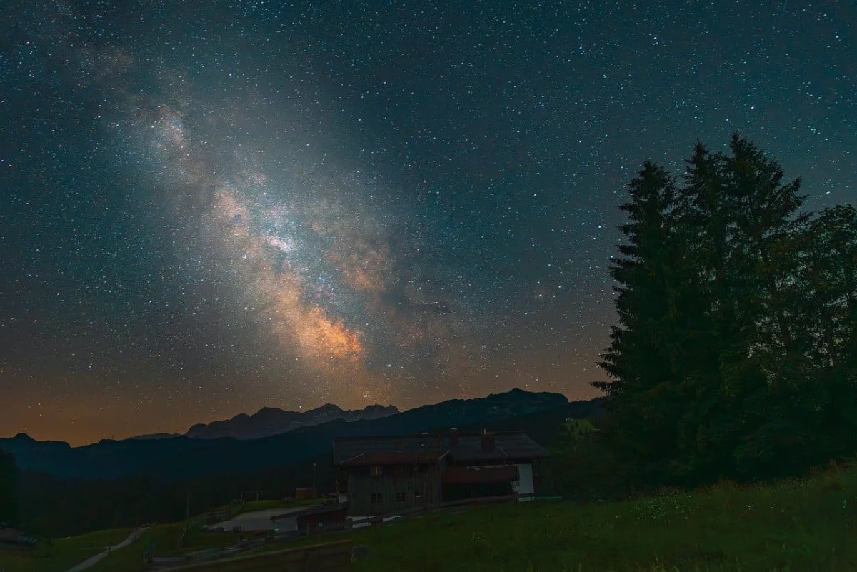 Vista de la Vía Láctea iluminando el cielo nocturno sobre un paisaje montañoso con árboles