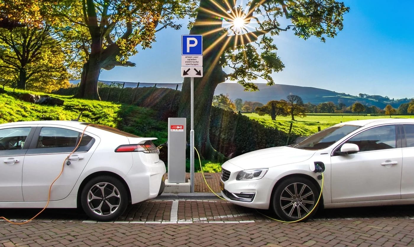 Dos coches eléctricos blancos cargando en una estación al aire libre con paisaje verde de fondo