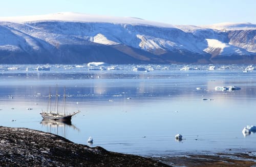Velero navegando entre hielo flotante frente a montañas nevadas en Groenlandia