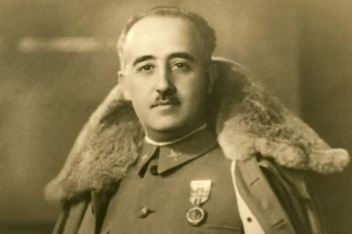 Retrato histórico en blanco y negro de Francisco Franco con uniforme militar y capa con cuello de piel, posando de pie en actitud solemne