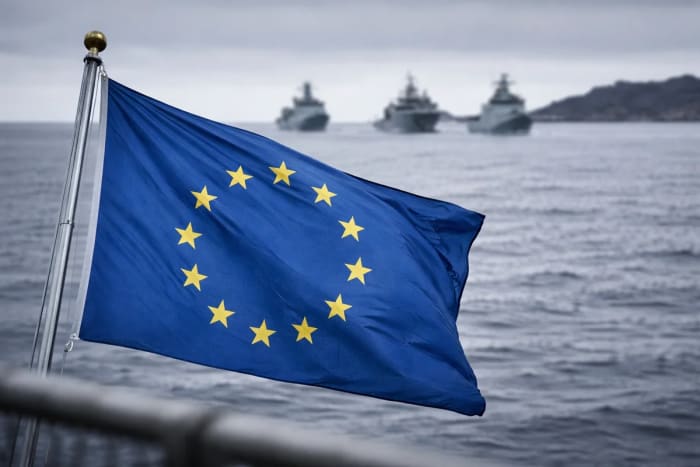 Bandera de la Unión Europea en primer plano con buques militares desenfocados en el mar al fondo