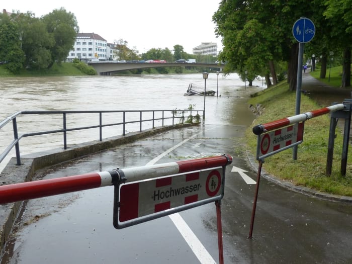 Inundación del río con paso peatonal anegado y señal de “Hochwasser” cerrando el acceso