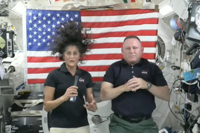 Astronautas en microgravedad dentro de una nave espacial con bandera de Estados Unidos al fondo