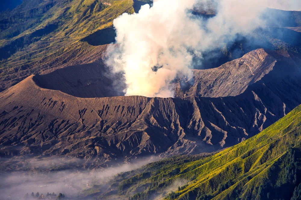 Vista aérea de un volcán humeante con flancos erosionados y vegetación en las laderas bajo un cielo claro.