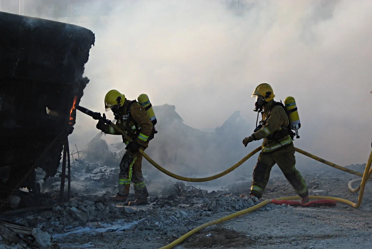 Dos bomberos en acción apagando un incendio entre escombros, usando mangueras de agua y equipo de respiración autónoma en un ambiente lleno de humo.