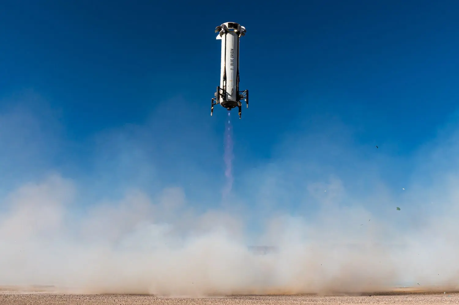 El cohete New Shepard de Blue Origin aterrizando de manera controlada en el sitio de lanzamiento, levantando una nube de polvo bajo un cielo despejado.