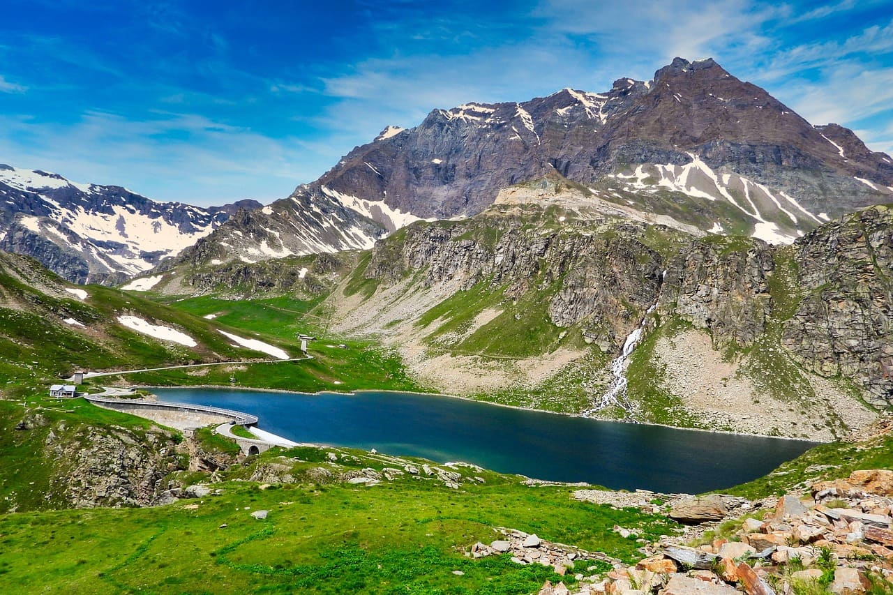 Embalse de alta montaña rodeado por cumbres nevadas y formaciones rocosas en los Alpes, bajo un cielo azul despejado.